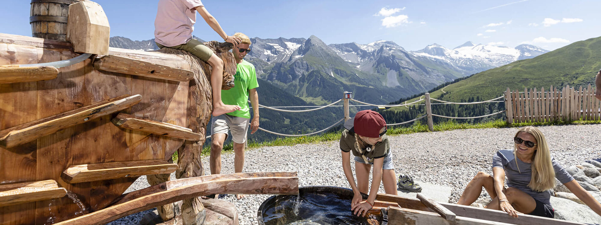 Eine Familie genießt einen sonnigen Tag in den Bergen, während Kinder an einem Wasserspielzeug spielen und lachen.