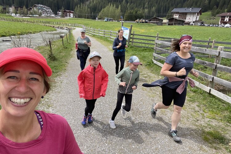 Eine Gruppe von Frauen und Mädchen joggt fröhlich auf einem malerischen Weg in den Bergen, umgeben von grüner Natur.