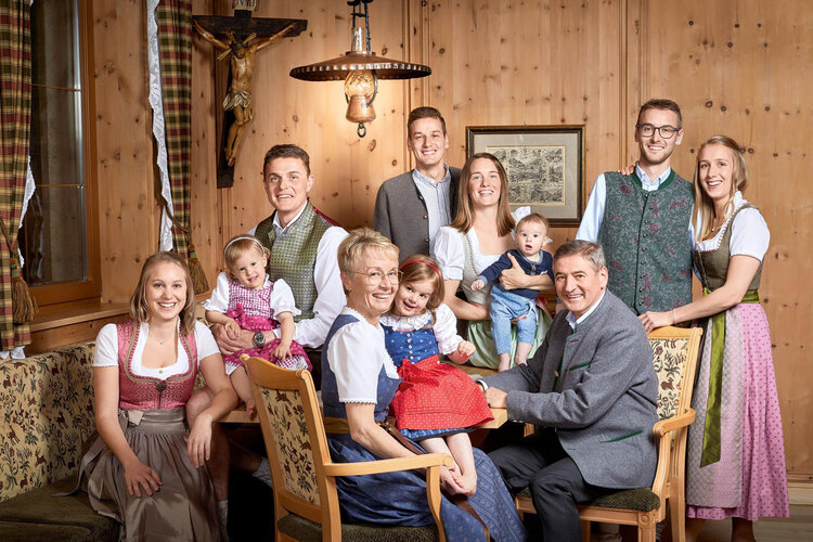 Eine fröhliche Familie posiert in traditioneller Tracht in einem gemütlichen Holzraum mit einem Kreuz im Hintergrund.