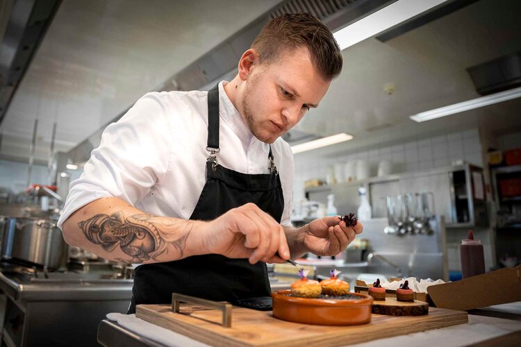 Der Koch in der weißen Uniform zeigt Präzision beim Anrichten von Desserts auf einem Holzbrett in einer hellen, modernen Küche.
