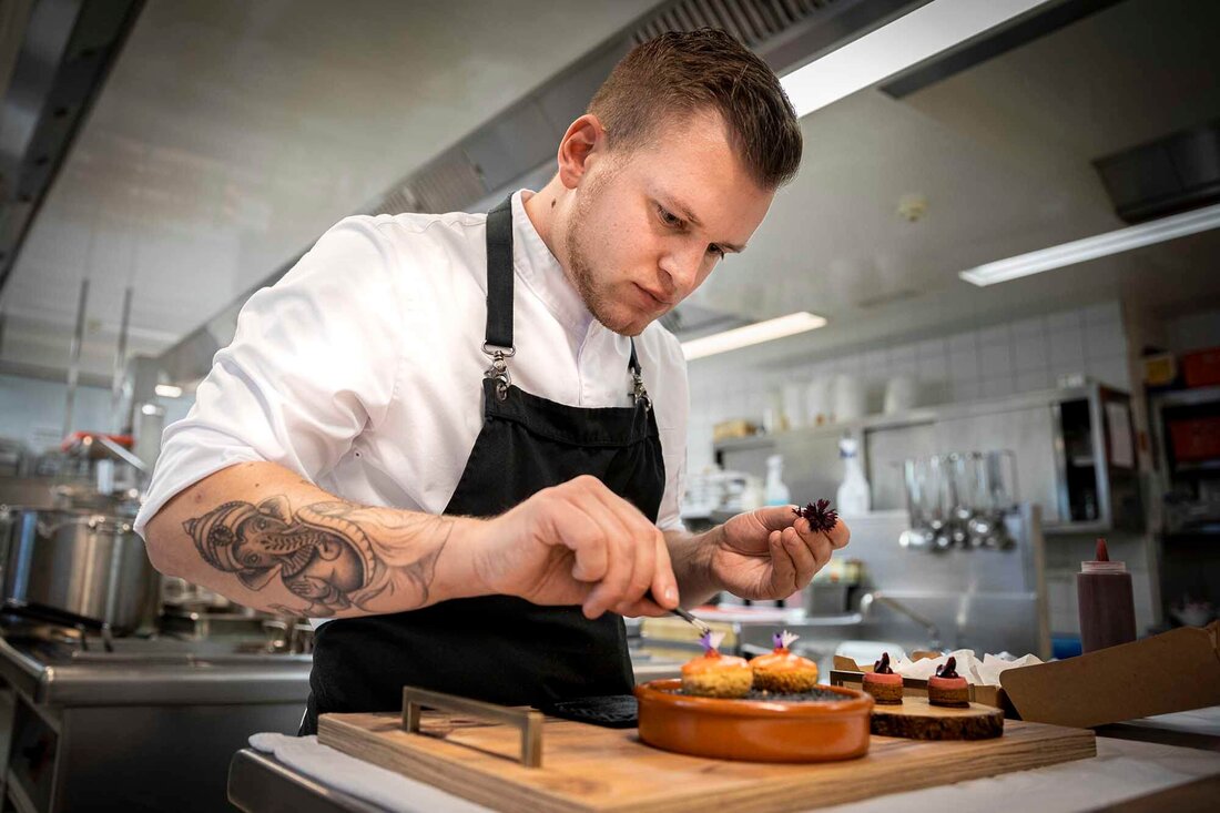 Der Koch in der weißen Uniform zeigt Präzision beim Anrichten von Desserts auf einem Holzbrett in einer hellen, modernen Küche.