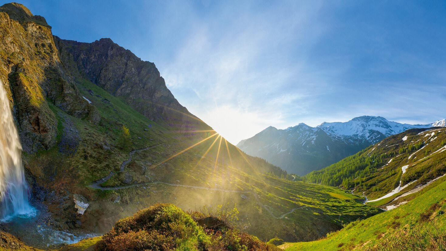 Die Sonne strahlt durch die Wolken und beleuchtet den Wasserfall, während die Landschaft in sattem Grün erblüht.