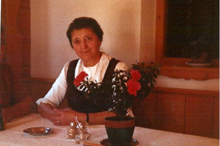 Die Frau in traditioneller Tracht lächelt sanft, während sie an einem Tisch mit einer blühenden Pflanze und Geschirr sitzt.