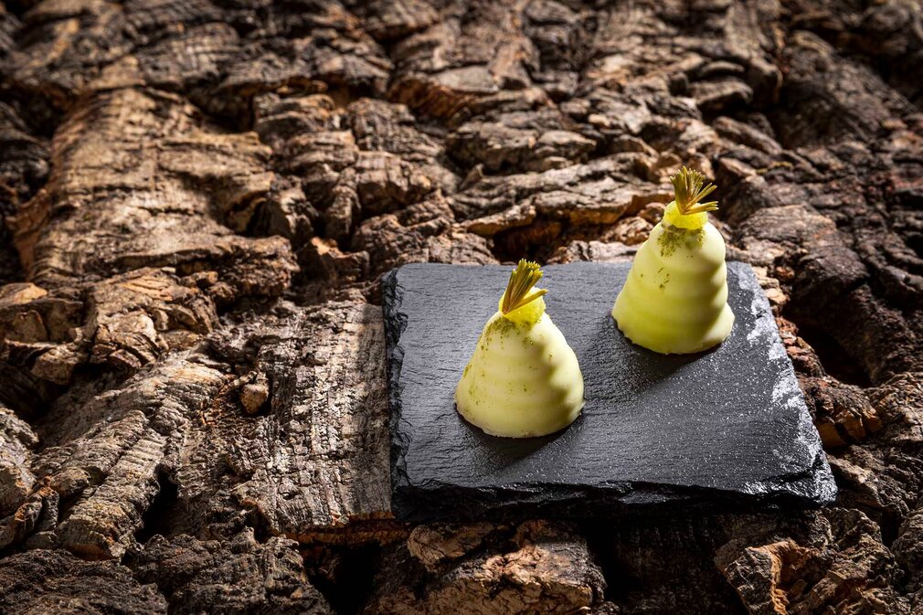 Zwei kleine, birnenförmige Desserts mit einer grünen Garnitur stehen auf einem schwarzen Teller, vor einer natürlichen Holztextur.