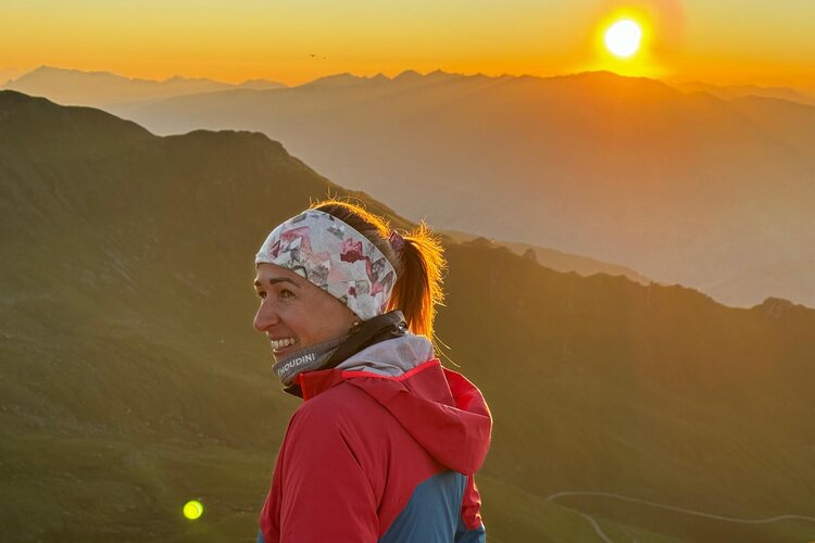 Eine lächelnde Frau mit einem bunten Stirnband genießt den Sonnenuntergang über den Bergen.
