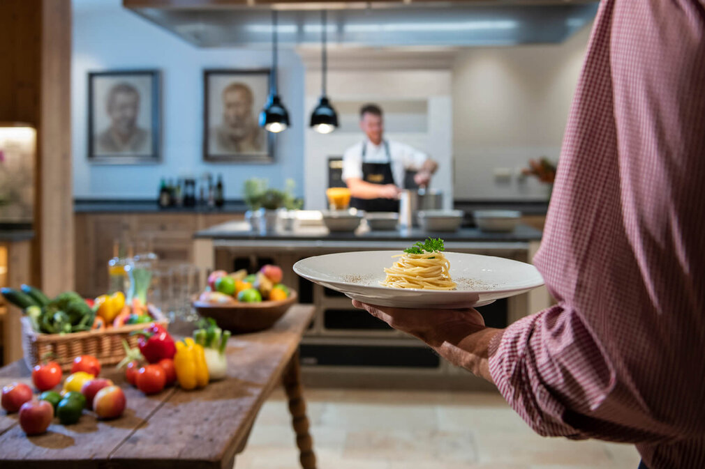In einer hellen Küche wird ein Teller mit Spaghetti von einem Mann gehalten, während ein Koch im Hintergrund aktiv ist.