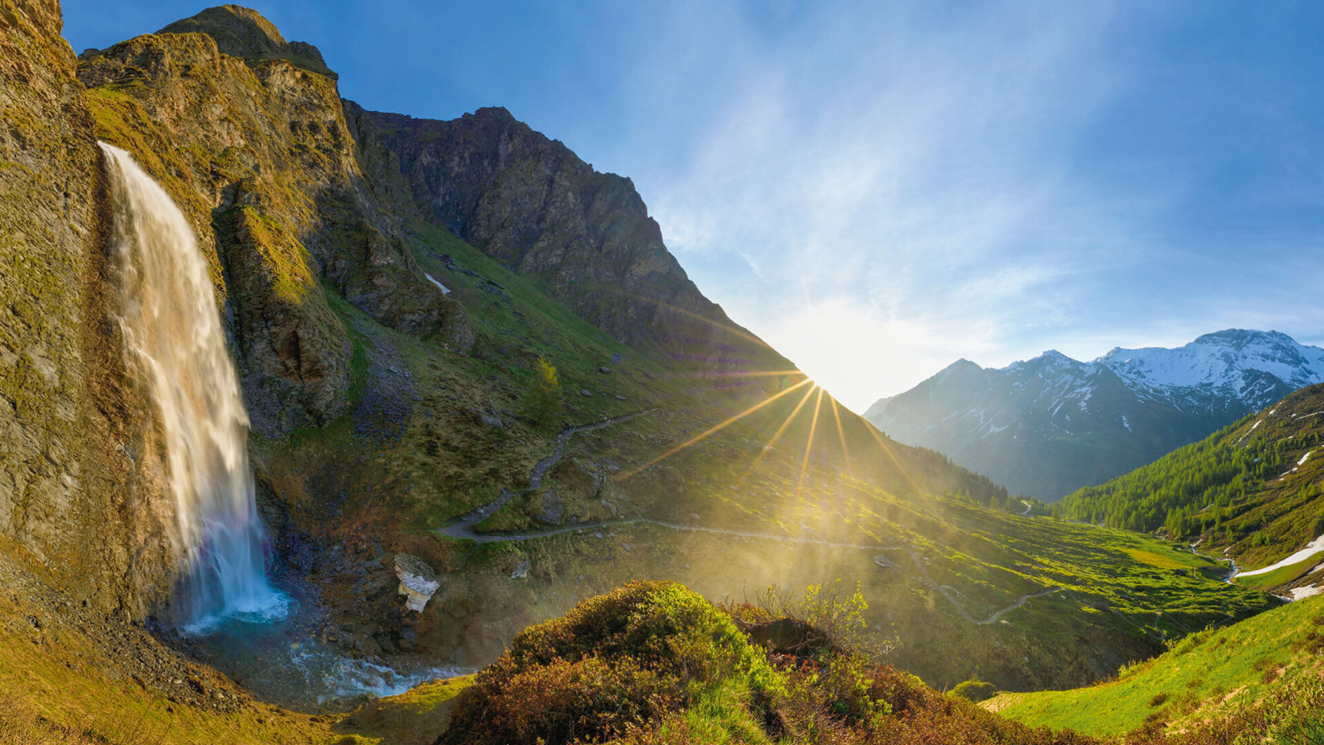 Ein majestätischer Wasserfall stürzt über eine steile Klippe, umgeben von üppigem Grün und majestätischen Bergen im Hintergrund.