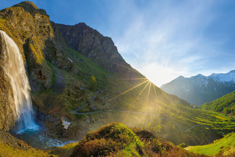 Ein majestätischer Wasserfall stürzt über eine steile Klippe, umgeben von üppigem Grün und majestätischen Bergen im Hintergrund.