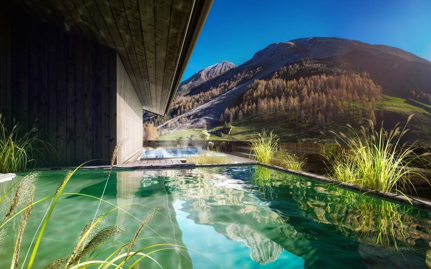Ein modernes Holzhaus mit einem Pool, umgeben von üppigem Grün und majestätischen Bergen im Hintergrund unter klarem Himmel.