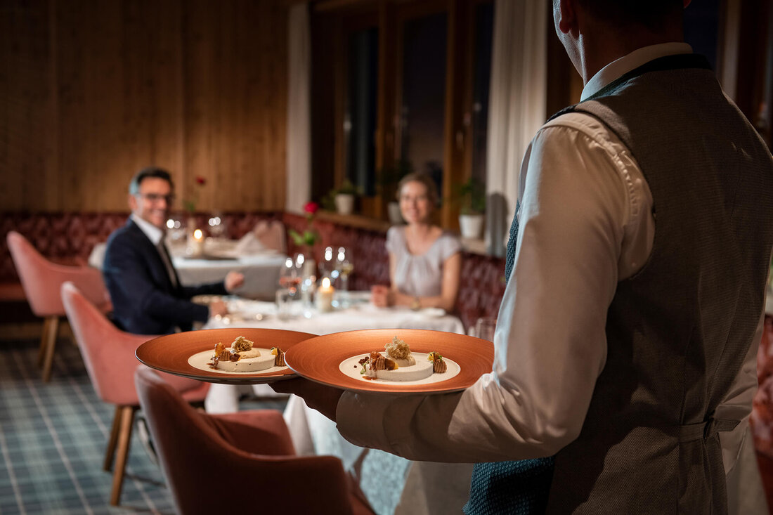Zwei Gäste genießen ein romantisches Dinner, während der Kellner ihnen exquisite Gerichte in einem warmen, holzverkleideten Raum bringt.