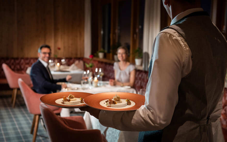 Zwei Gäste genießen ein romantisches Dinner, während der Kellner ihnen exquisite Gerichte in einem warmen, holzverkleideten Raum bringt.