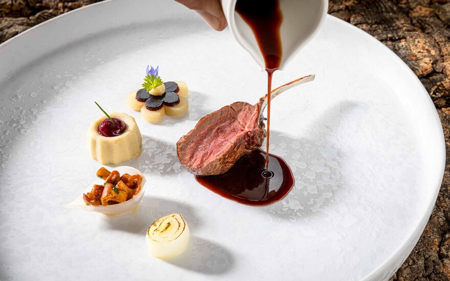 Ein zartes Lammkotelett wird mit einer dunklen Sauce übergossen, umgeben von kunstvoll angerichteten Beilagen.