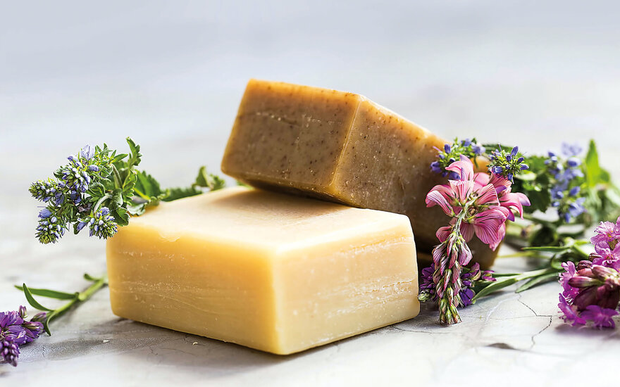 Zwei Seifenstücke liegen auf einem hellen Untergrund, umgeben von frischen, bunten Blumen und Kräutern.
