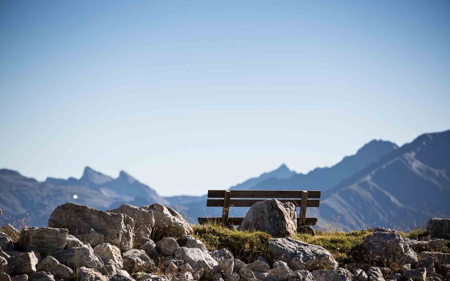 Eine Holzbank steht einsam auf einem Hügel, umgeben von großen Steinen und mit majestätischen Bergen im Hintergrund.