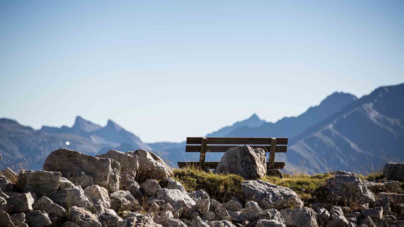 Eine Holzbank steht einsam auf einem Hügel, umgeben von großen Steinen und mit majestätischen Bergen im Hintergrund.
