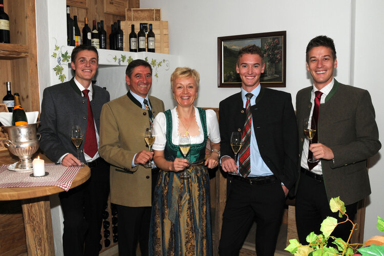 Fünf Personen in traditioneller Tracht stehen lächelnd in einem gemütlichen Raum mit Weinflaschen im Hintergrund.