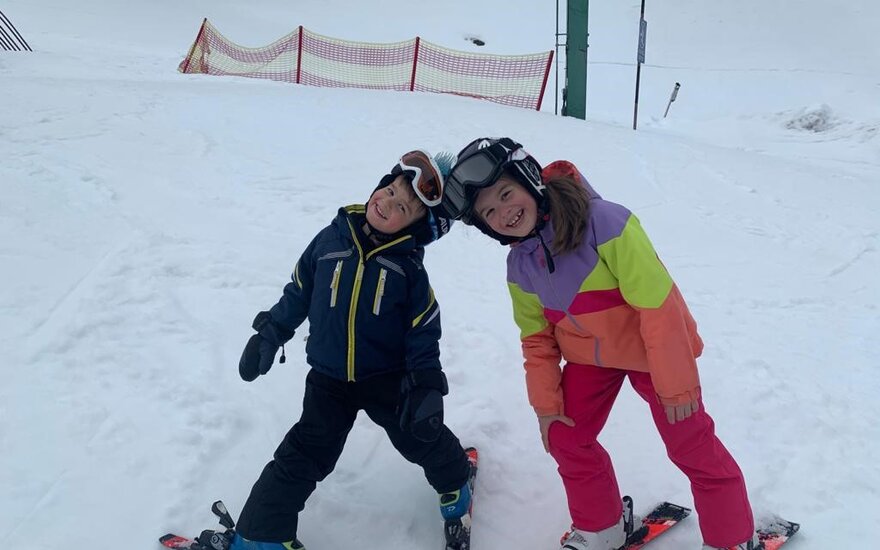 Zwei fröhliche Kinder stehen auf Skiern im Schnee, lächeln und posieren für die Kamera, umgeben von einer winterlichen Landschaft.