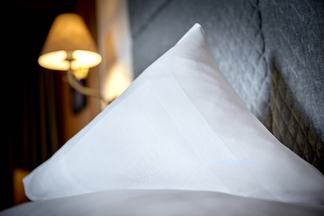 Ein elegantes, weißes Kissen liegt auf einem Bett, während eine sanfte Lampe im Hintergrund warmes Licht ausstrahlt.