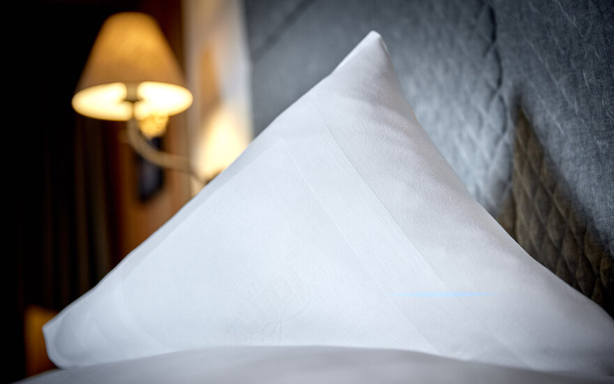 Ein elegantes, weißes Kissen liegt auf einem Bett, während eine sanfte Lampe im Hintergrund warmes Licht ausstrahlt.