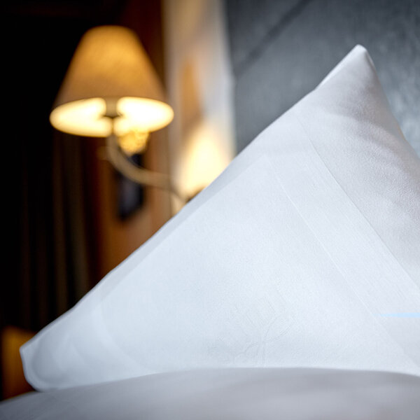 Ein elegantes, weißes Kissen liegt auf einem Bett, während eine sanfte Lampe im Hintergrund warmes Licht ausstrahlt.