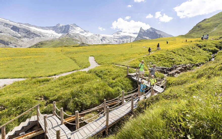Eine Gruppe von Wanderern überquert eine Holzbrücke in einer blühenden, grünen Landschaft mit Bergen im Hintergrund.