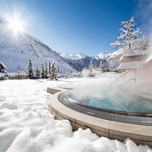 Ein Whirlpool im Freien, umgeben von schneebedeckten Bergen und Tannen, während die Sonne strahlt und Dampf aufsteigt.