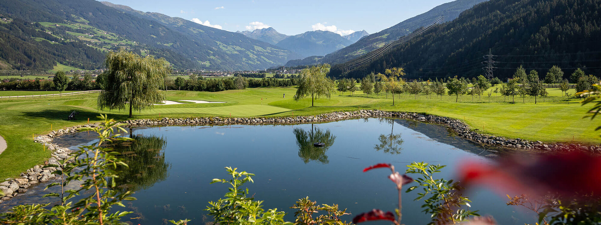 Eine malerische Golfanlage mit einem ruhigen Teich, umgeben von sanften Hügeln und majestätischen Bergen im Hintergrund.