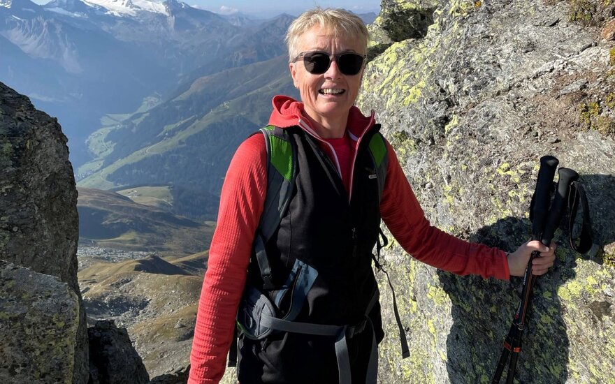 Eine fröhliche Wandererin steht an einem felsigen Hang mit atemberaubendem Bergblick im Hintergrund.