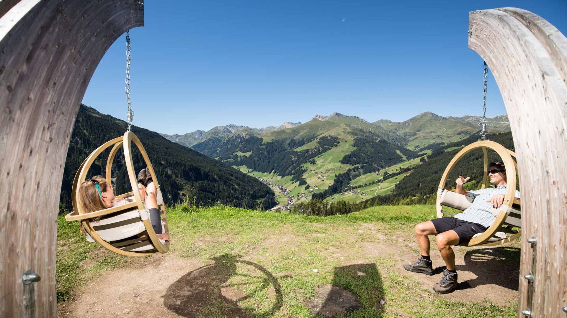 Ein Mann und eine Frau genießen die Aussicht in schwingenden Sesseln, umgeben von majestätischen Bergen und klarem Himmel.