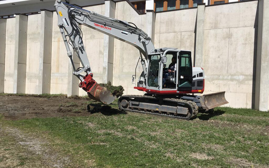Ein Bagger hebt Erde an, während ein Fahrer im Führerhaus sitzt, umgeben von einer Baustelle mit Betonwänden.