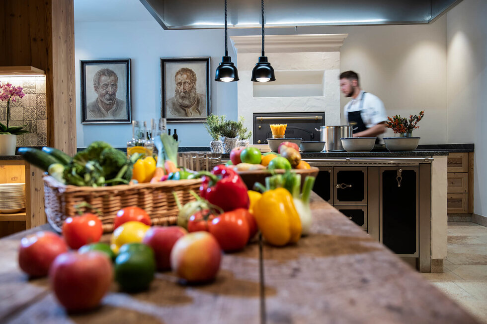 Ein gut gefüllter Tisch mit frischem Gemüse und Obst, während ein Koch im Hintergrund beschäftigt ist.