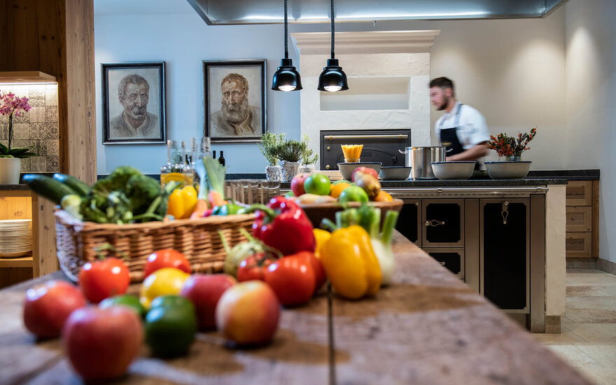 Ein gut gefüllter Tisch mit frischem Gemüse und Obst, während ein Koch im Hintergrund beschäftigt ist.