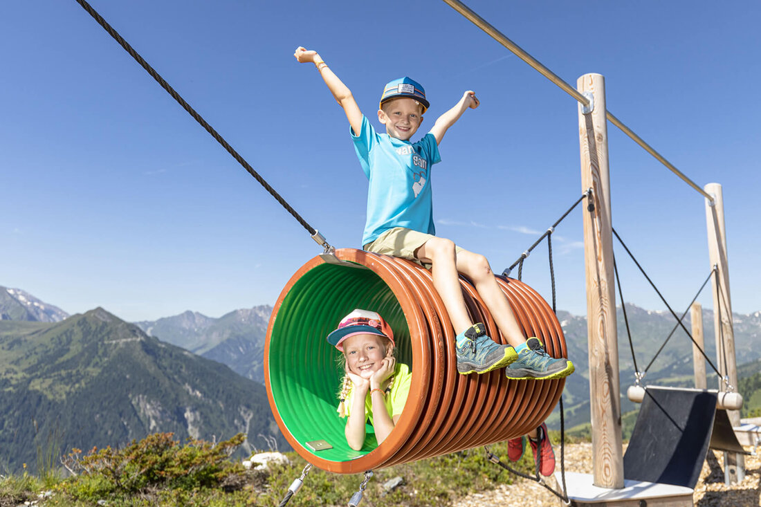 Zwei fröhliche Kinder spielen in einem bunten Tunnel auf einem Spielplatz mit Bergblick, voller Freude und Energie.