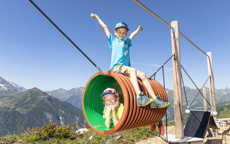 Zwei fröhliche Kinder spielen in einem bunten Tunnel auf einem Spielplatz mit Bergblick, voller Freude und Energie.