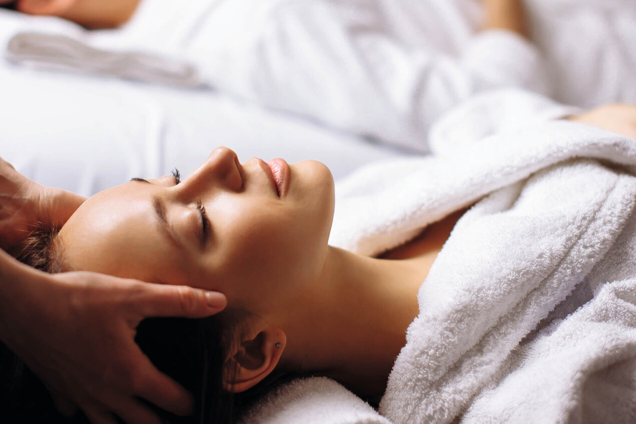 Eine Frau genießt eine entspannende Kopfmassage, während sie in einem weißen Bademantel auf einer Liege liegt.