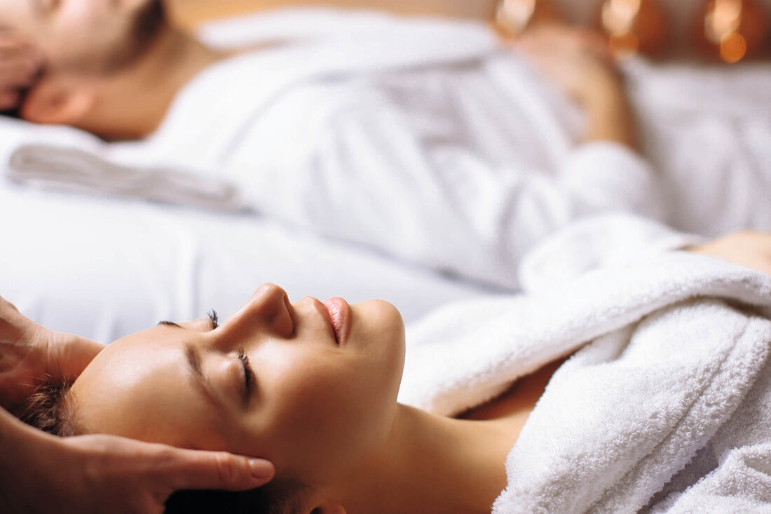 Eine Frau genießt eine entspannende Kopfmassage, während sie in einem weißen Bademantel auf einer Liege liegt.
