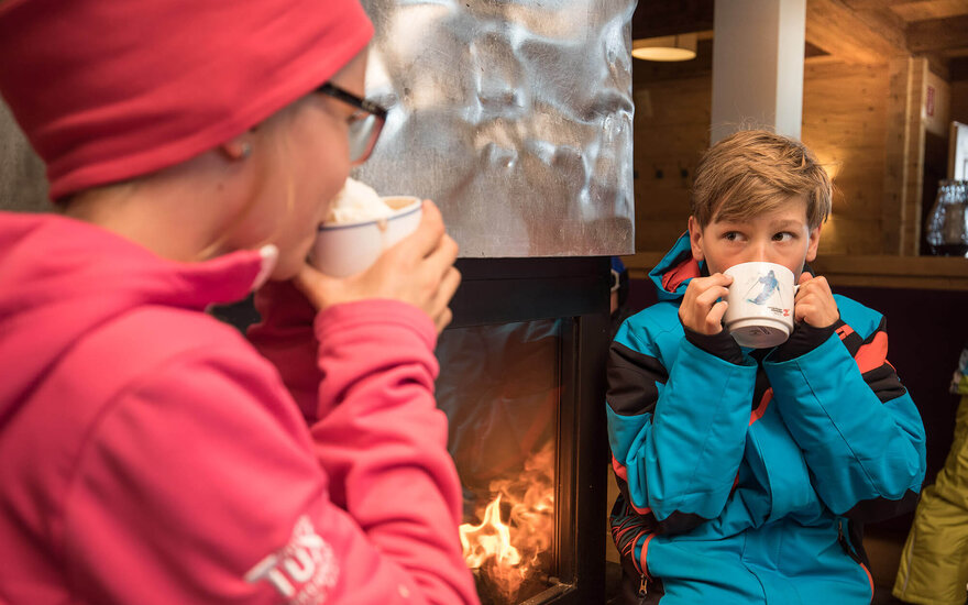 Ein Junge in blauer Winterjacke hält eine Tasse und schaut neugierig, während ein Mädchen in rosa im Hintergrund trinkt.