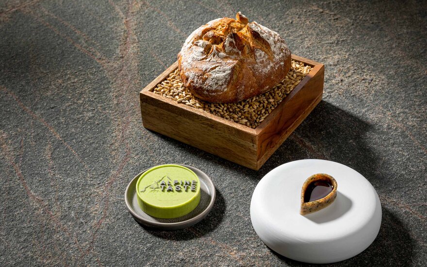 Ein rustikales Brot liegt in einer Holzschale, umgeben von goldenen Körnern, auf einer dunklen Steinoberfläche.