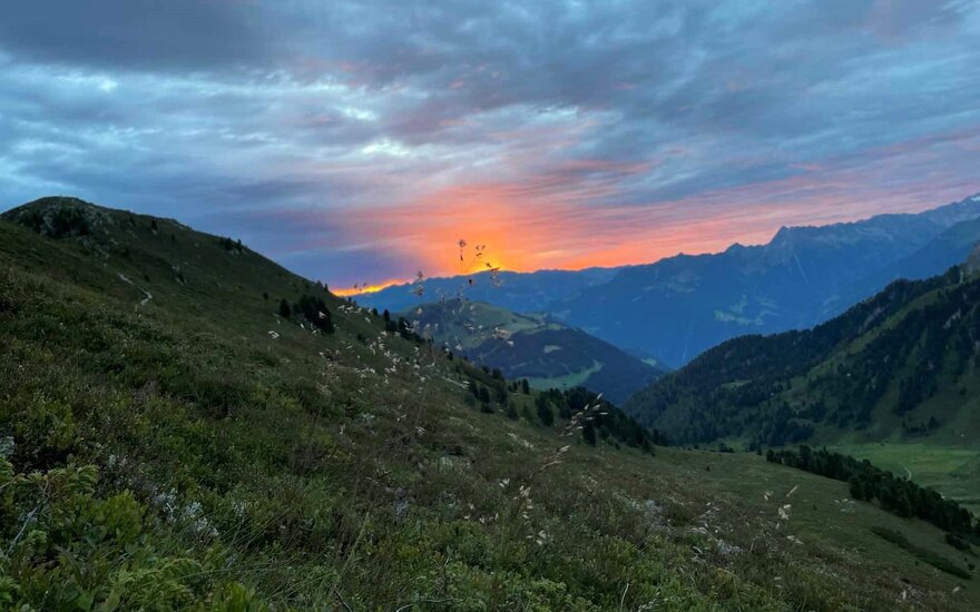 Ein atemberaubender Sonnenuntergang über den Bergen, mit sanften Hügeln und üppigem Grün im Vordergrund.
