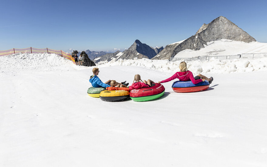 Eine Gruppe von vier Personen sitzt in bunten Reifen auf einer schneebedeckten Fläche, umgeben von majestätischen Bergen.