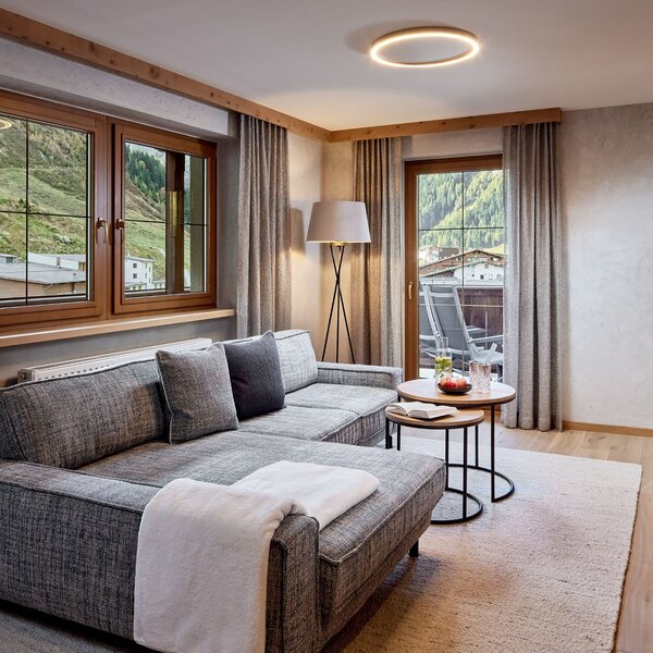 Ein modernes Wohnzimmer mit einem grauen Sofa, einem kleinen Tisch und großen Fenstern, die auf eine grüne Berglandschaft blicken.