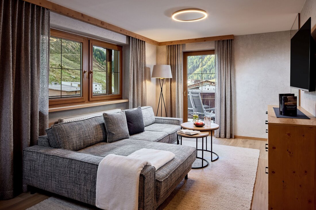 Ein modernes Wohnzimmer mit einem grauen Sofa, einem kleinen Tisch und großen Fenstern, die auf eine grüne Berglandschaft blicken.