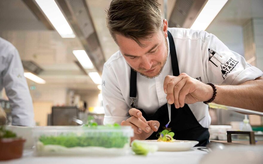 Ein Koch verziert sorgfältig einen Teller mit frischen Kräutern und essbaren Blumen in einer modernen Küche.