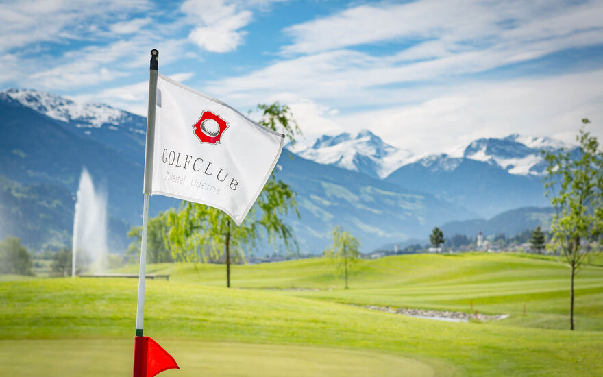 Eine weiße Fahne mit dem Logo des Golfclubs Zillertal weht sanft im Wind, umgeben von einer malerischen Berglandschaft.