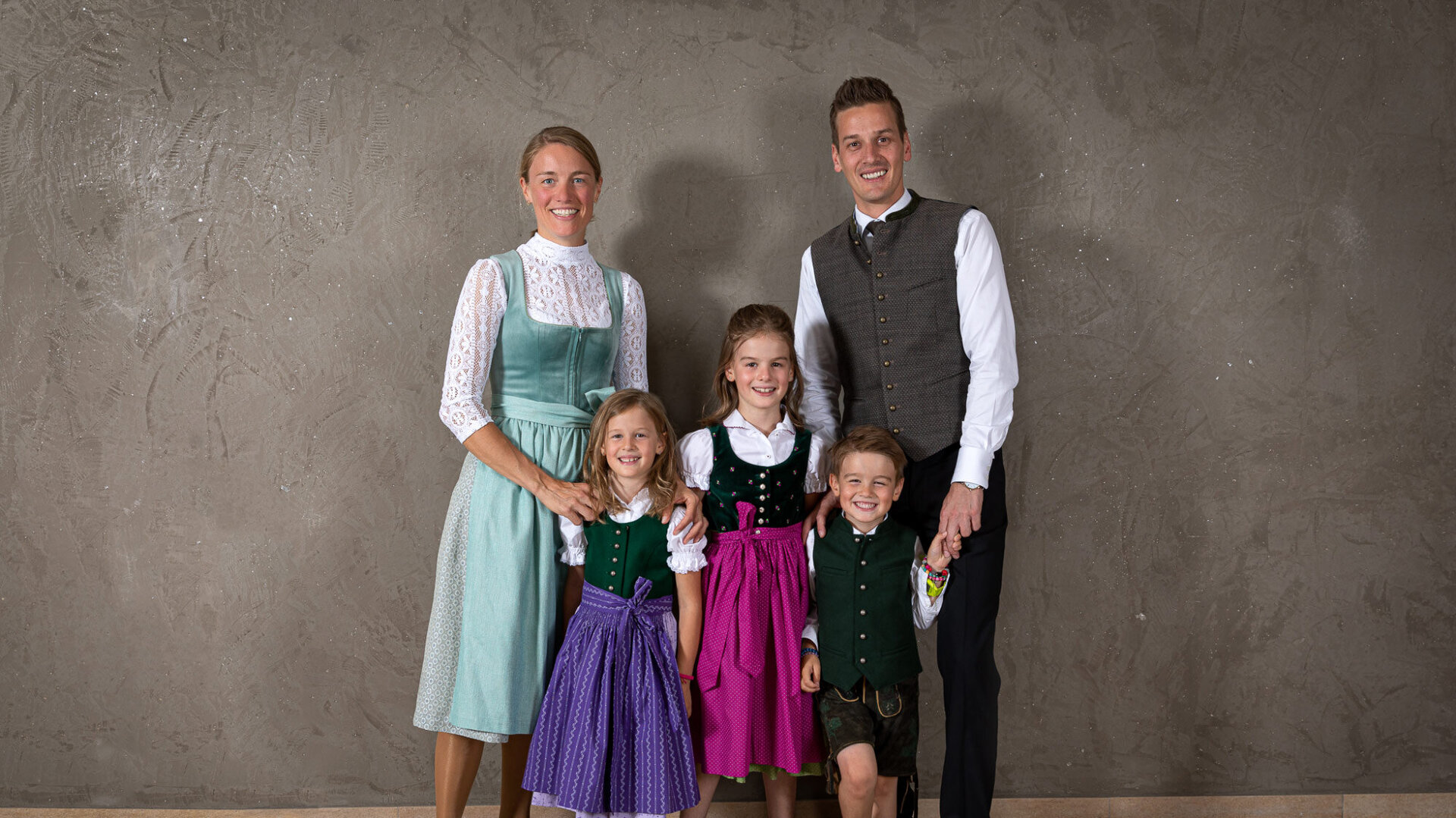 Eine fröhliche Familie posiert in traditioneller Tracht vor einer neutralen Wand, die Kinder lächeln begeistert.