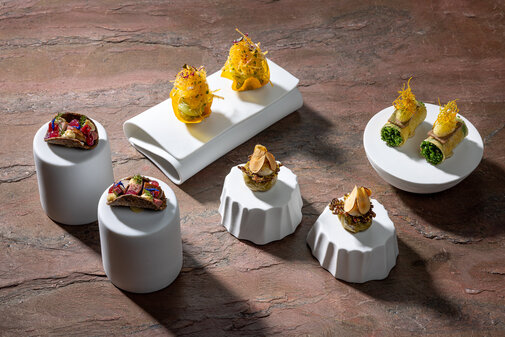 Drei verschiedene kleine Gerichte auf eleganten weißen Unterlagen, die mit frischen Zutaten und bunten Kräutern dekoriert sind.