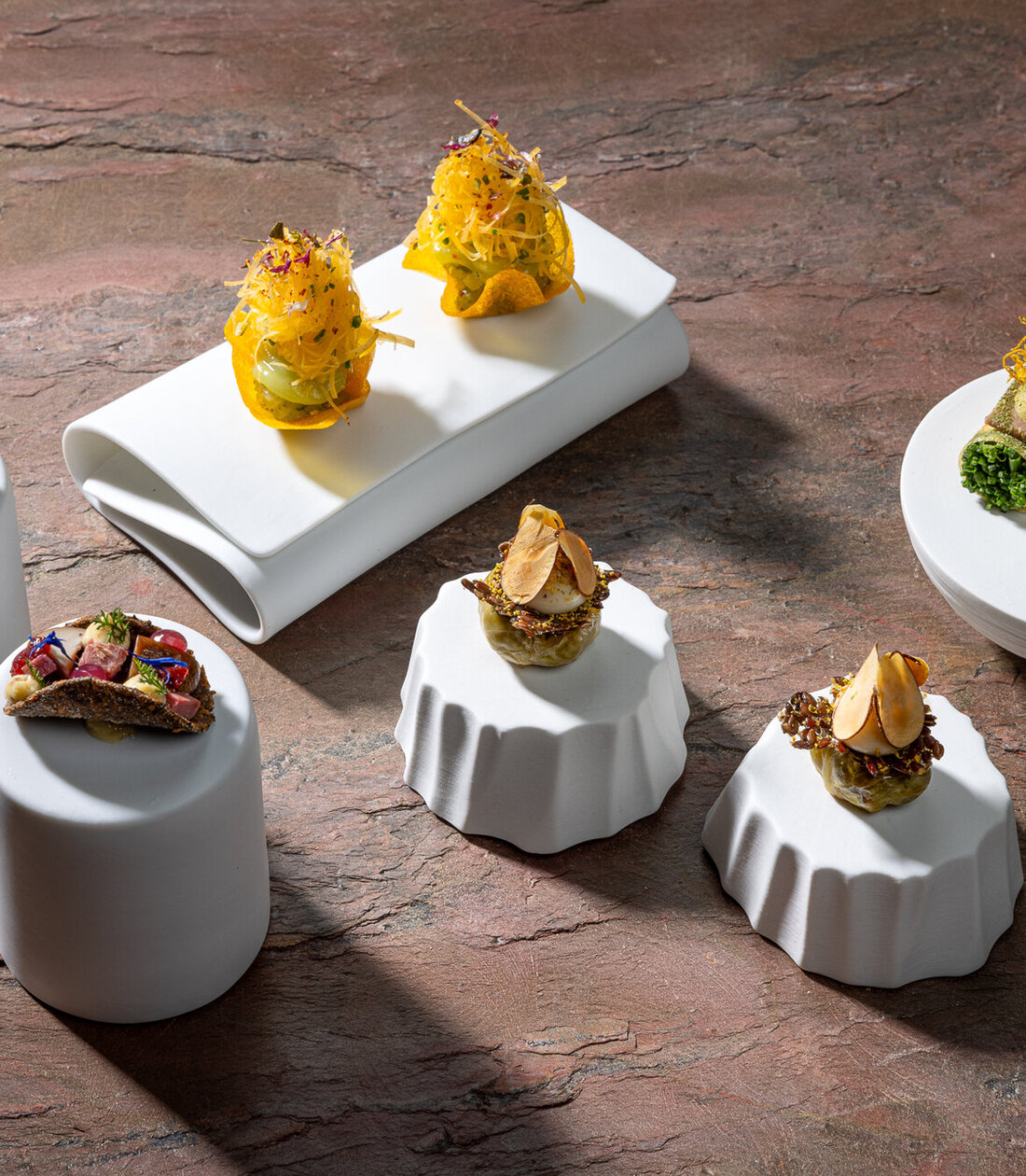 Drei verschiedene kleine Gerichte auf eleganten weißen Unterlagen, die mit frischen Zutaten und bunten Kräutern dekoriert sind.