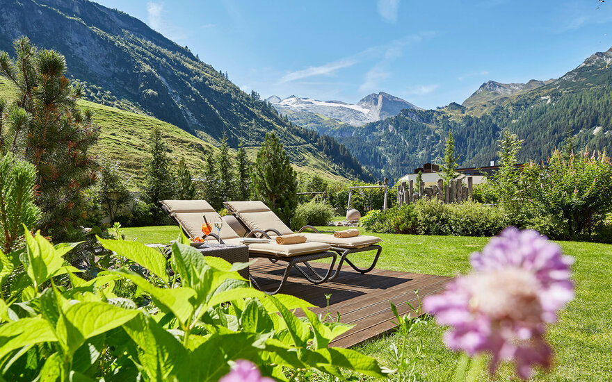 Zwei Liegestühle stehen auf einer Holzterrasse, umgeben von üppigem Grün und Blick auf majestätische Berge im Hintergrund.