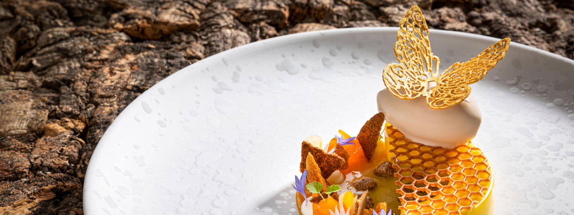 Ein kunstvoll angerichtetes Dessert mit einer goldenen Schmetterlingsdekoration und bunten essbaren Blüten auf einem weißen Teller.