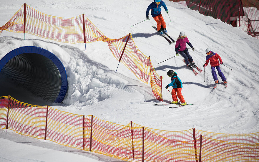 Vier Skifahrer, darunter Kinder, fahren fröhlich einen schneebedeckten Hang hinunter, umgeben von Sicherheitsnetzen.